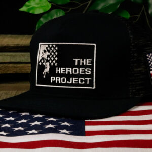 black hat with The Heroes Project embroidered logo sitting on a wooden table with a plant behind and American flag in front
