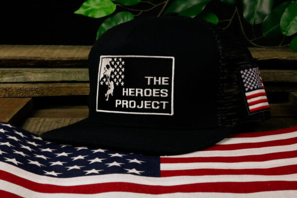 black hat with The Heroes Project embroidered logo sitting on a wooden table with a plant behind and American flag in front