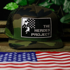 camo hat with The Heroes Project embroidered logo sitting on a wooden table with a plant behind and American flag in front
