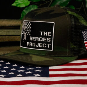 camo hat with The Heroes Project embroidered logo sitting on a wooden table with a plant behind and American flag in front